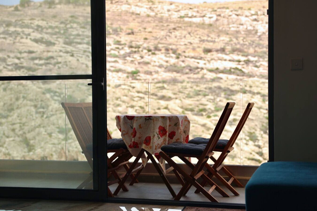 A room with a large window offering a view of a rocky landscape. There is a table with a floral tablecloth and two wooden chairs with cushions.