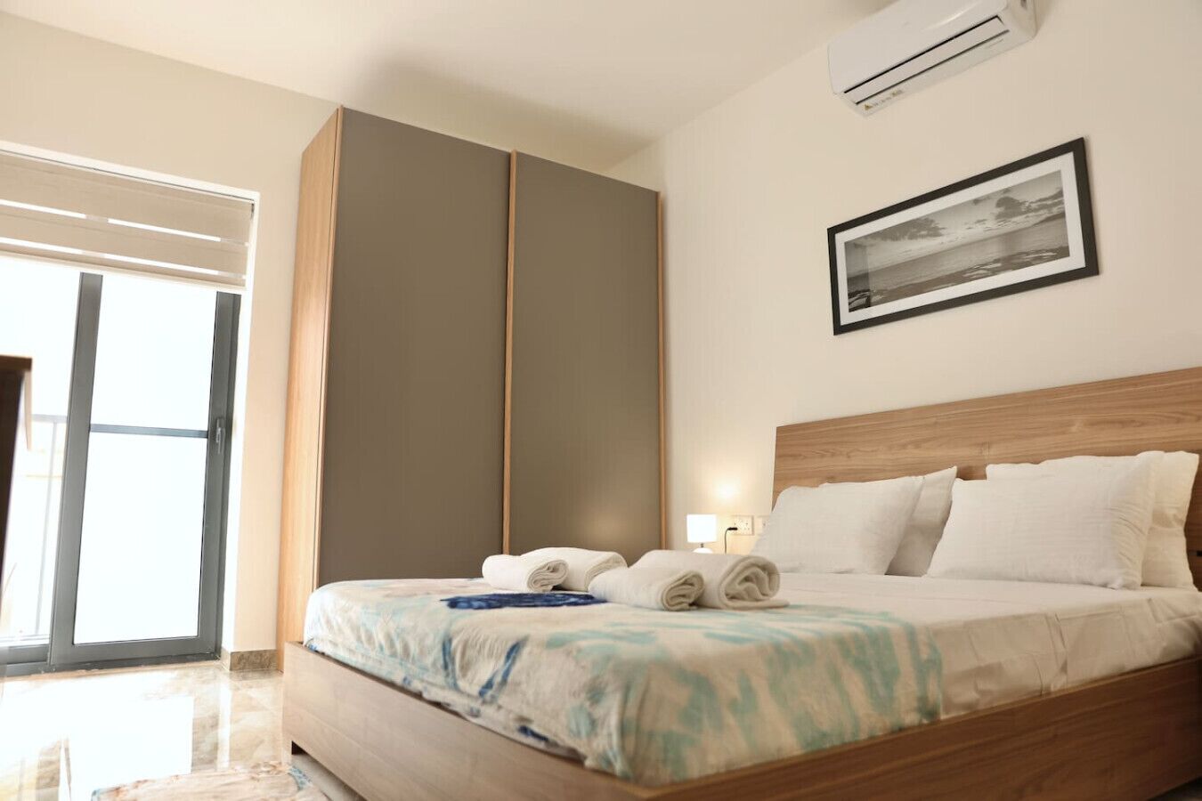 Une chambre d’hôtel avec un grand lit, une armoire, un tableau encadré au mur et une fenêtre avec des stores.