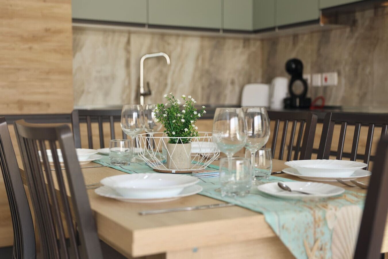 Una mesa de comedor dispuesta con platos blancos, cubiertos y copas de vino, con una planta en maceta como centro.