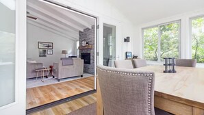 A modern living room with a fireplace, a dining area with a wooden table, and a large window offering a view of trees. - Pet-Friendly Oasis w/ Sunroom – Steps to Beach! (Douglas)