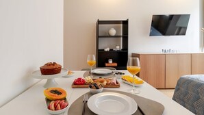 A breakfast setup with a variety of foods and drinks on a table, a television mounted on the wall, and a bookshelf in the background. - Central & Cozy Studio by HostWise (Vila Nova de Gaia)