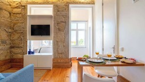 A cozy room with a stone wall, a flat-screen TV mounted above a white cabinet, a dining table with breakfast items, and a blue chair. - Cozy Studio with Workspace by HostWise (Porto)