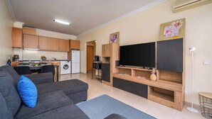 A modern living room with a grey sofa, a flat-screen TV, wooden shelves, and a kitchen area with cabinets and appliances.