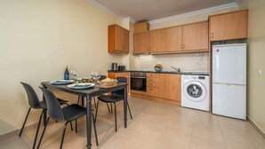 A compact kitchen with wooden cabinets, a built-in oven, a washing machine, and a dining area with a table and chairs.