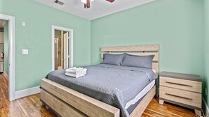 A bedroom with a wooden bed, a nightstand, a ceiling fan, and a door leading to a bathroom. - Sunny Hidden Retreat C – Ocean Town Gem (Galveston)