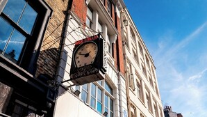 A traditional clock with "BAR ITALIA" and "ESPRESSO" signs, attached to a building with large windows and a brick facade.