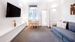 Una sala de estar moderna con un televisor de pantalla plana montado en la pared, una zona de comedor con mesa y sillas, un sofá gris con cojines y un ventanal que proporciona luz natural.