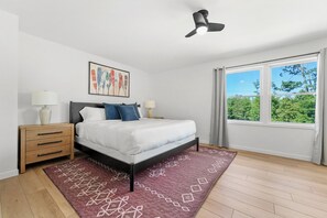 A bedroom with a bed, bedside tables, a ceiling fan, a window with curtains, and a view of greenery outside.