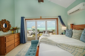 Un dormitorio con una cama grande, un aparador de madera, un espejo y vistas a la playa a través de una puerta corrediza de vidrio.