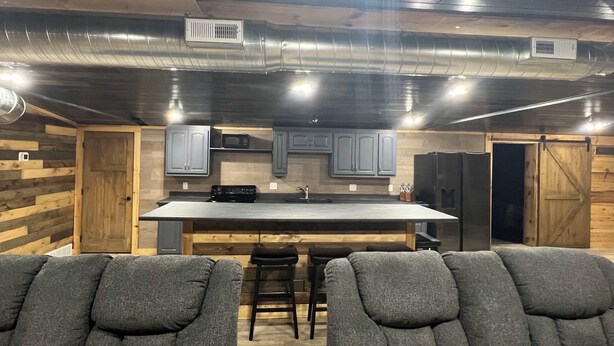 A modern kitchen with a central island, grey cabinets, and a black refrigerator. There are two grey upholstered chairs in the foreground. - Boar's Tusk Lodge (Winner)