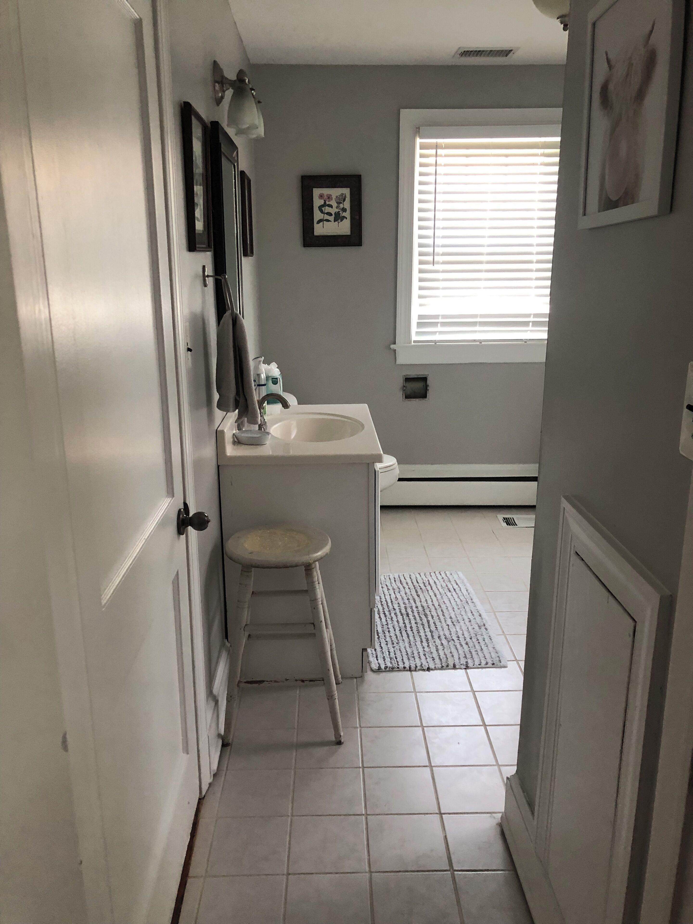 A narrow bathroom with a white sink, a stool, and a framed picture on the wall.