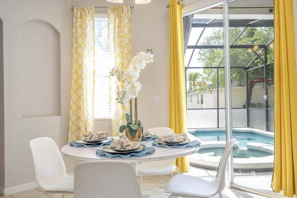 Un coin repas avec une table ronde, des chaises blanches, des rideaux jaunes et une vue sur une piscine.