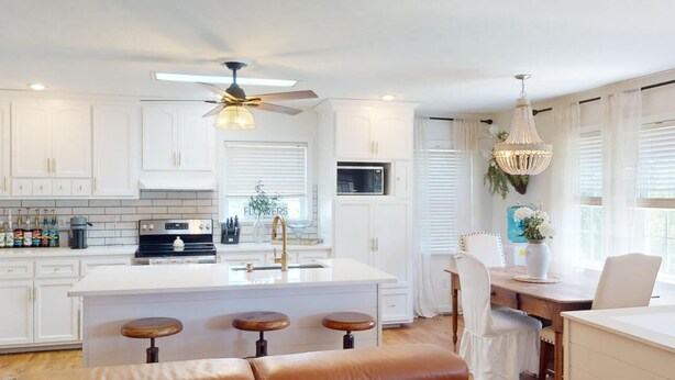 A modern kitchen with white cabinetry, a central island with stools, a ceiling fan, and a dining area with a chandelier.
