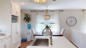A kitchen with white cabinetry, a stainless steel sink, a chandelier, a clock, and a window with curtains.