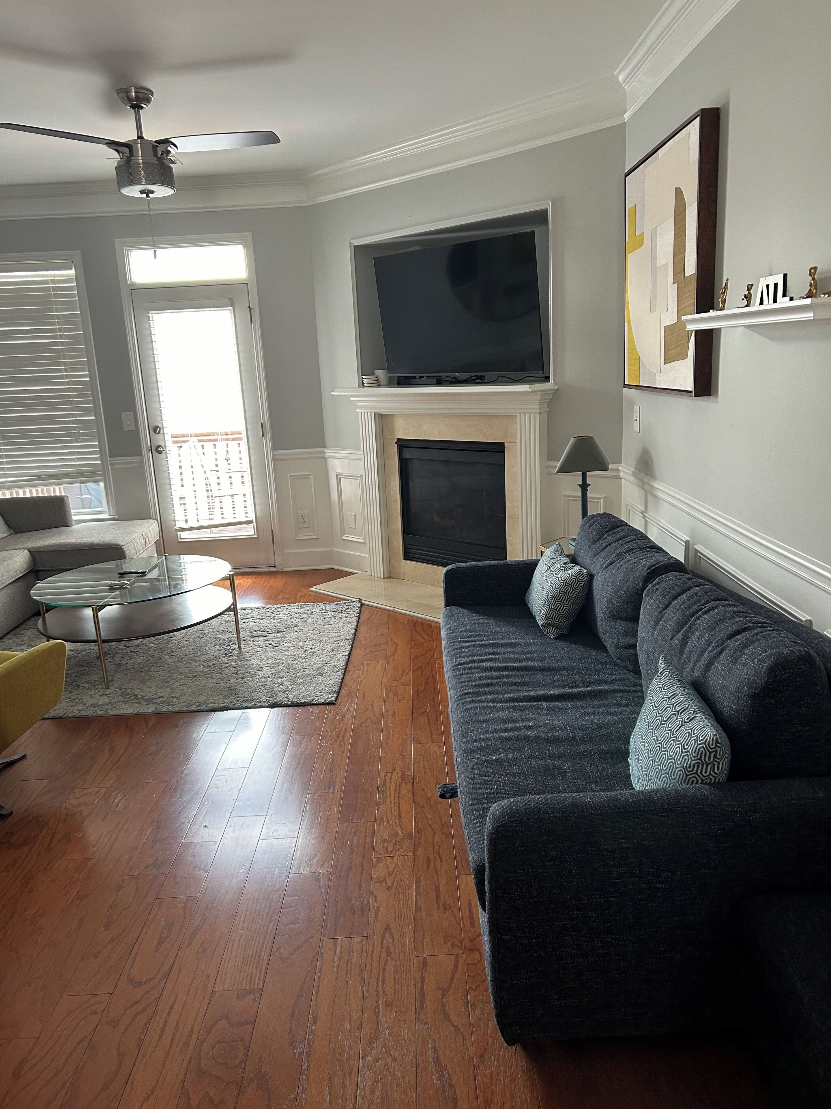 A living room with a dark-colored sofa, a flat-screen TV mounted above a fireplace, a ceiling fan, and a door leading to a balcony.