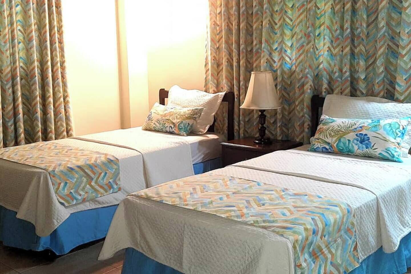 Two single beds with patterned bedspreads, a nightstand with a lamp, and curtains with a chevron pattern.