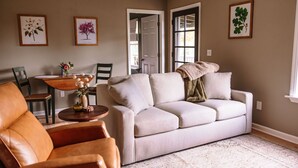 Una sala de estar con un sofá beige, un sillón de cuero marrón, una mesa de madera con un florero y grabados botánicos enmarcados en las paredes.