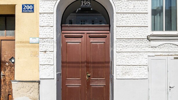 Dos puertas marrones con un picaporte de latón, una pared blanca con textura y un letrero azul con el número 198 sobre la puerta.