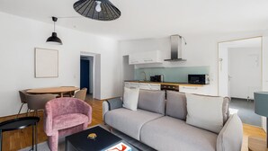 Una sala moderna con un sofá gris, un sillón rosa y una mesa de centro negra. Cuenta con una zona de cocina con armarios blancos y un microondas.