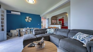A modern living room with a grey sectional sofa, a wooden coffee table, and a dining area with a blue wall and a decorative plate wall hanging. - Wave & Chill Apartment with garden 2 steps from the sea (Sainte-Luce)