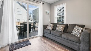 A living room with a gray sofa, patterned cushions, a sliding glass door leading to an outdoor patio, and a window with blinds.