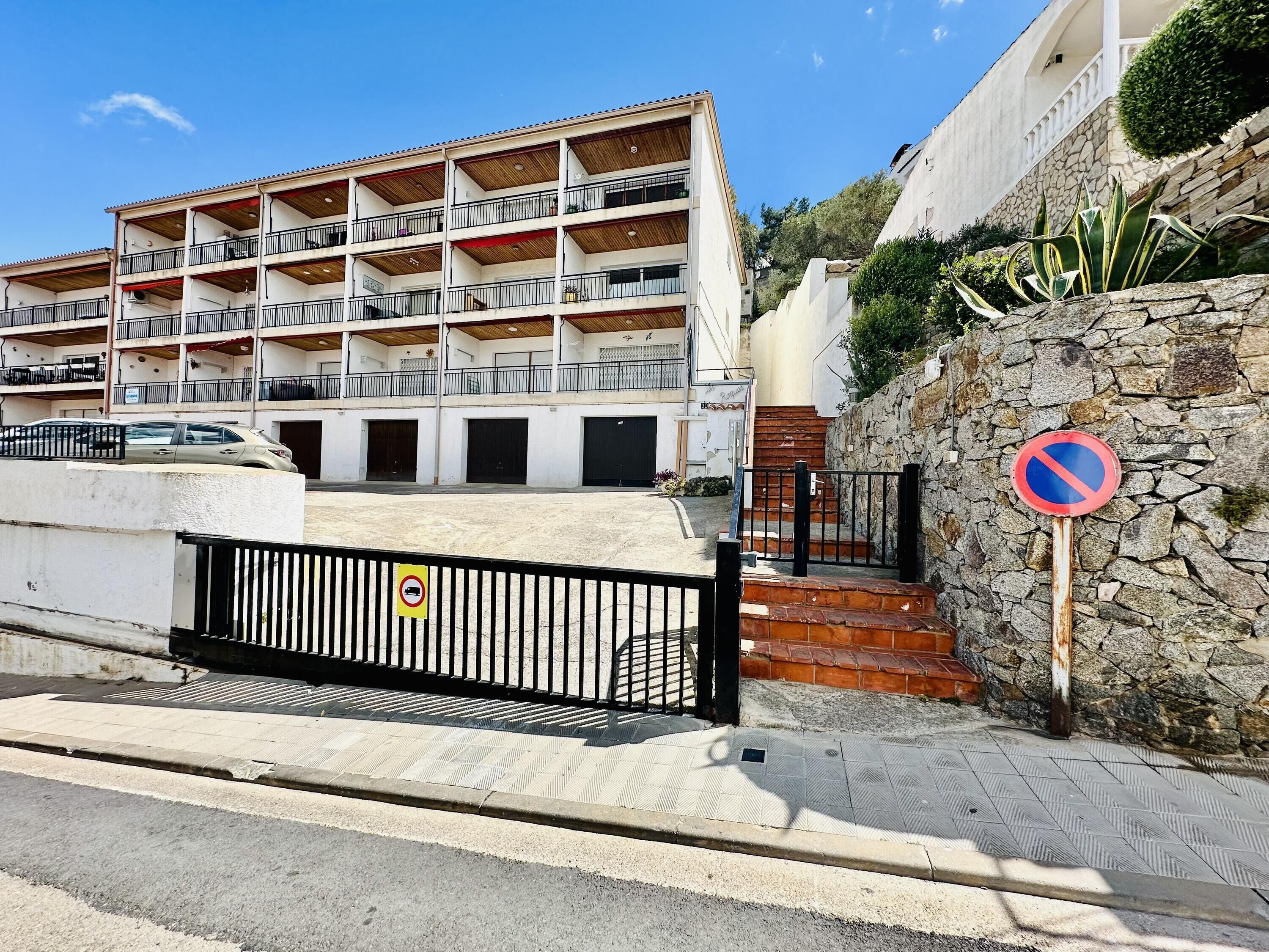 A multi-story building with balconies, a no parking sign, and a stone wall with plants.