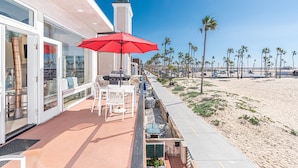 En patio ved stranden med rød parasol, udendørs møbler og udsigt til strand og palmer.