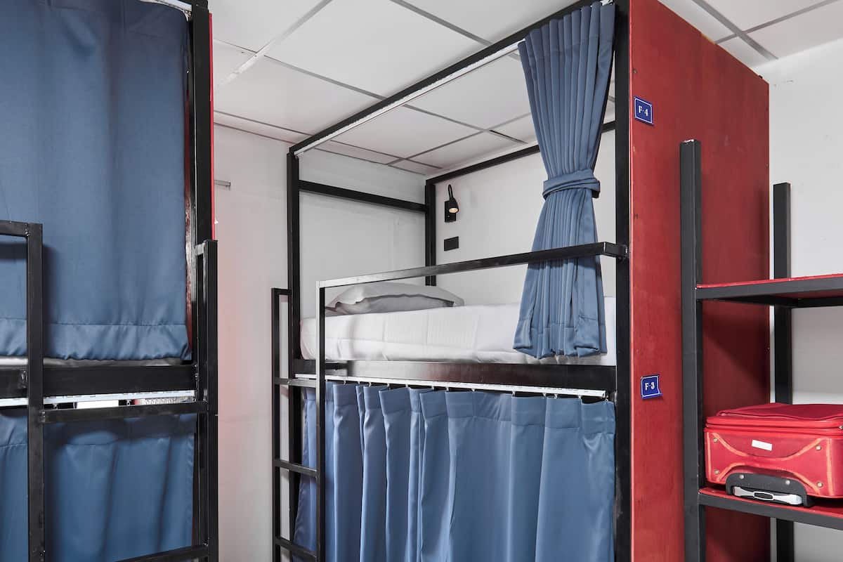 A bunk bed room with a red suitcase on a shelf, a blue curtain, and a white wall with a light switch.