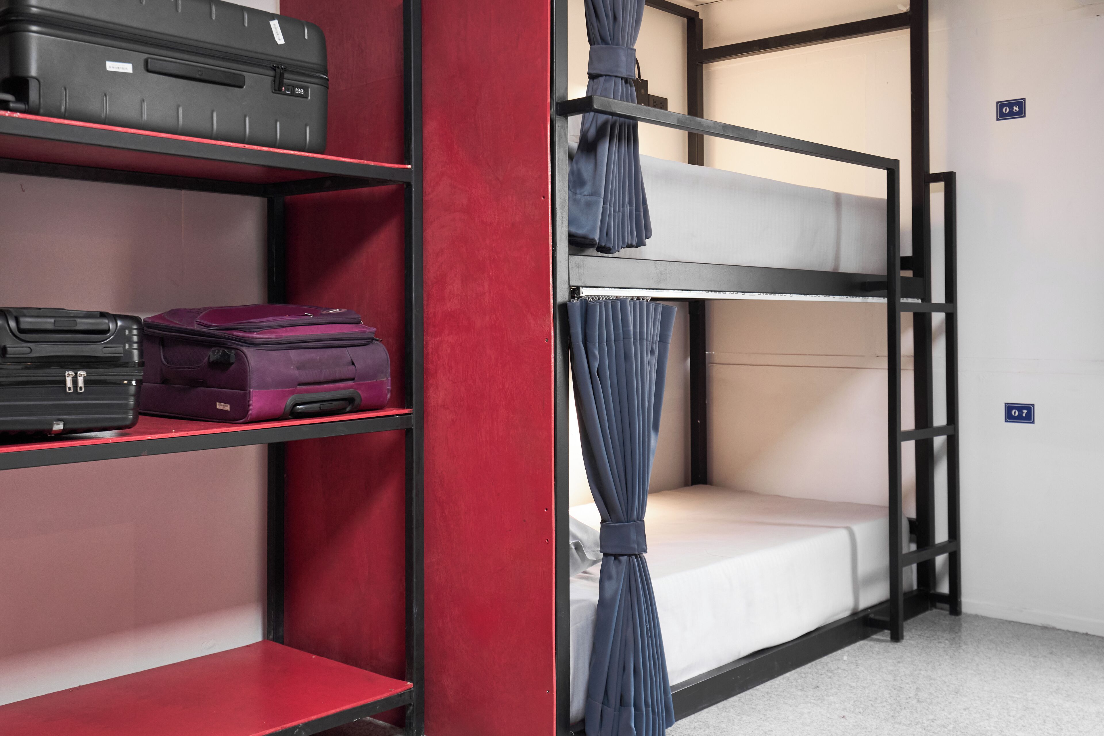 A bunk bed room with a red wall, a shelf with luggage, and a curtain.
