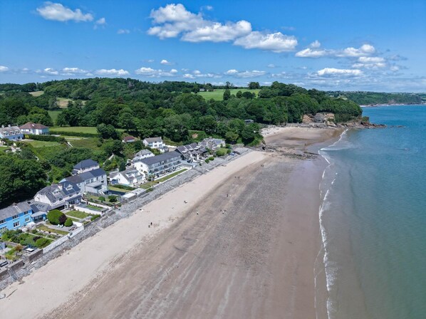 Une zone côtière avec une plage de sable, une rangée de maisons et une colline boisée en arrière-plan.
