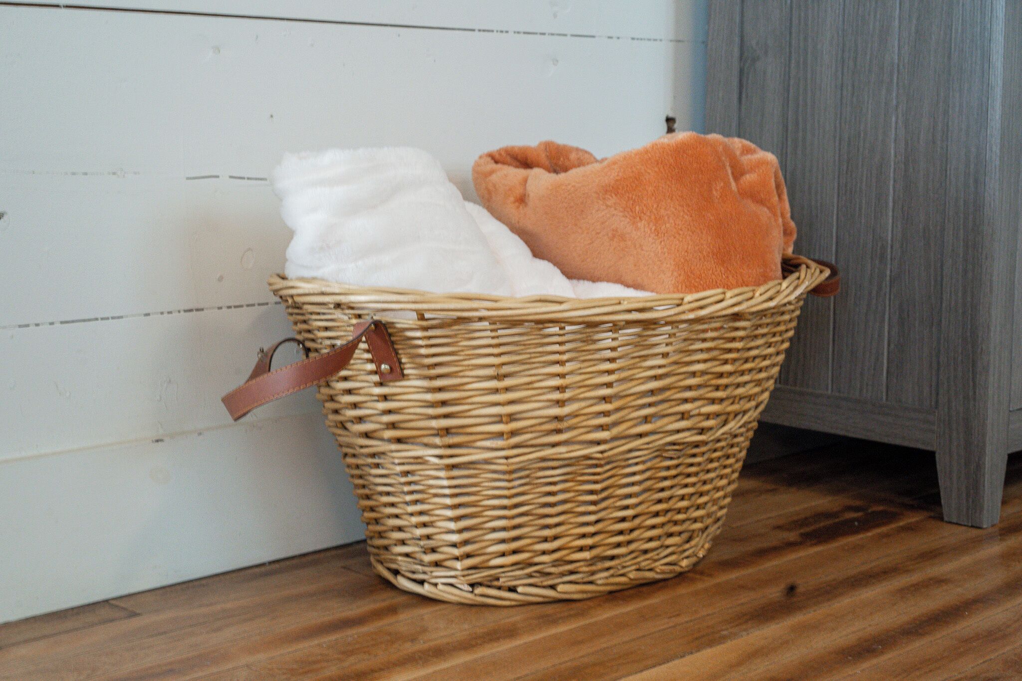 Een rieten mand met een bruin handvat op een houten vloer voor een witte muur.