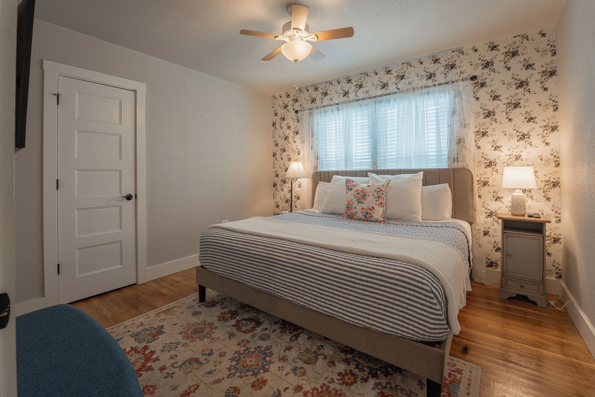 A bedroom with a bed, a ceiling fan, a window with blinds, a nightstand with a lamp, and a door.