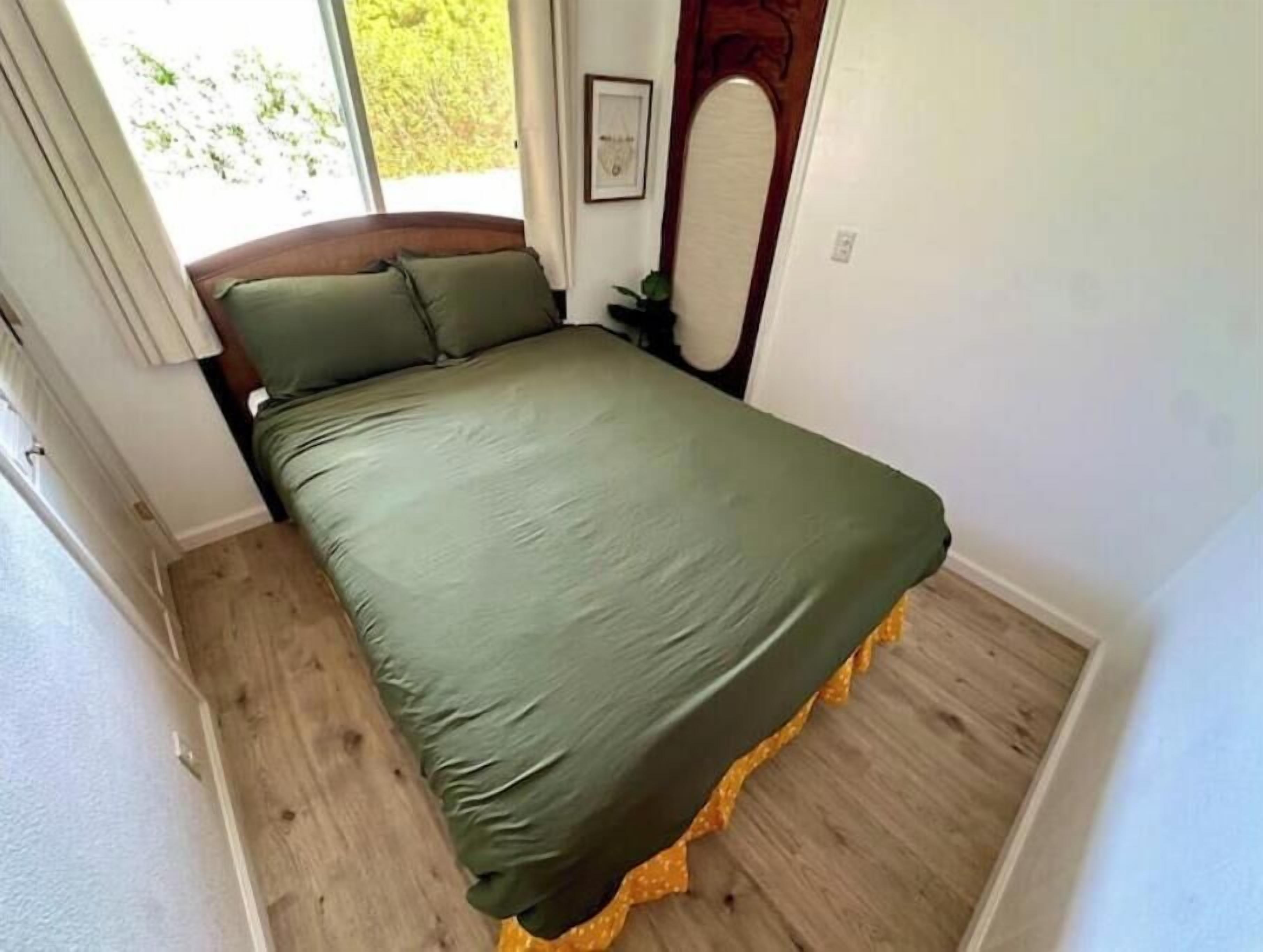 Un lit simple avec une couvre-lit verte, dans une pièce dont le sol est en bois et qui possède une fenêtre.