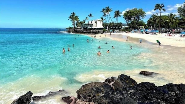 Une plage avec une eau turquoise et limpide, des gens qui se baignent et prennent des bains de soleil, et un bâtiment avec des palmiers en arri ère-plan.
