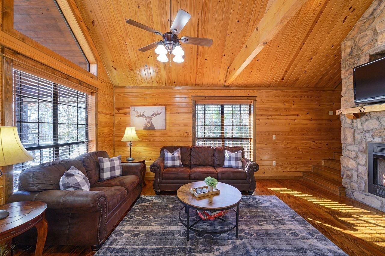 Interior acogedor de una cabaña de madera con chimenea, dos sofás de cuero, una mesa de centro y un ventilador de techo.