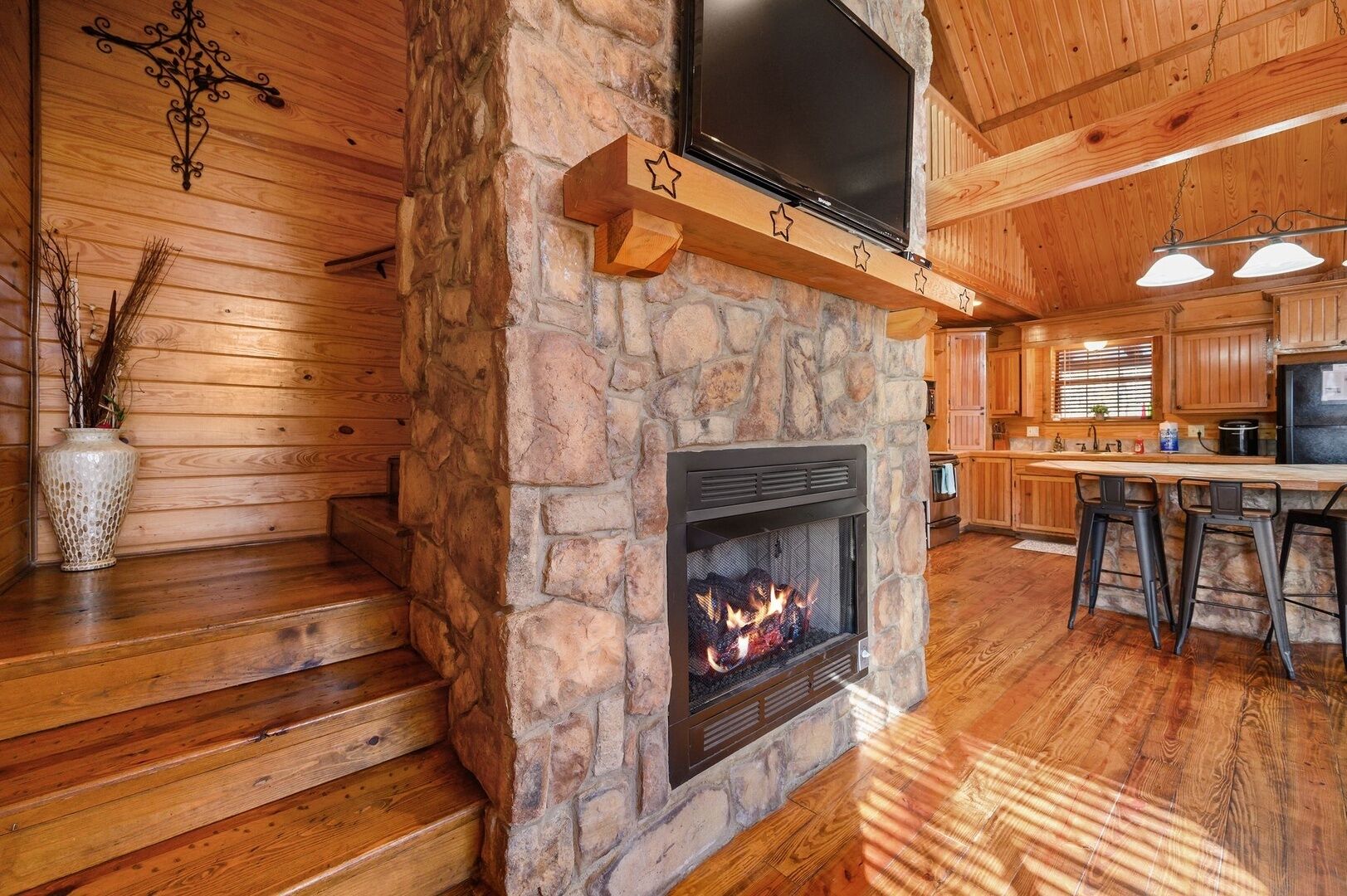 Ein Kamin aus Stein mit einem Fernseher darüber, Holzboden und ein Küchenbereich mit Barhocker.
