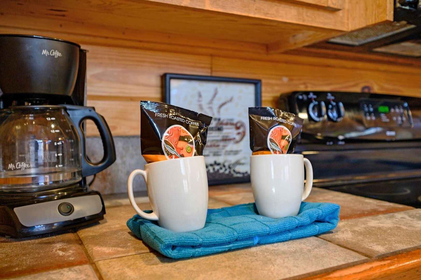 Un comptoir de cuisine avec une machine à café de la marque Mr. Coffee, deux tasses blanches contenant des dosettes de café et un torchon bleu.