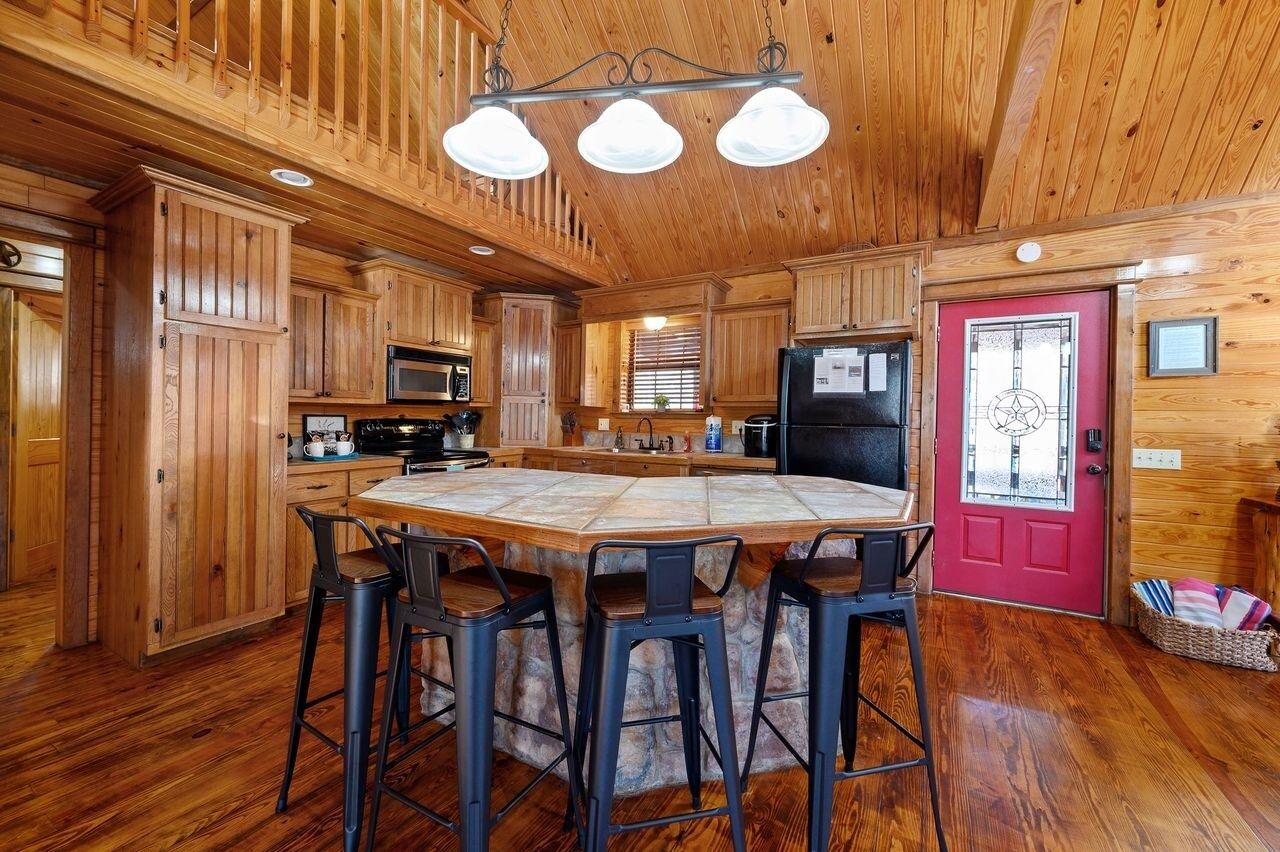 Interieur van een houten chalet met een eilandtafel in het midden, barstoelen, houten kasten, een rode deur en een hangende lamp.