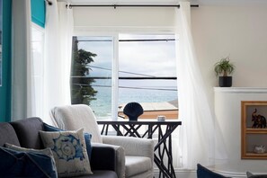 Uma sala de estar com vista para o mar, um sofá com almofadas estampadas, uma planta em vaso e uma estante com objetos decorativos.