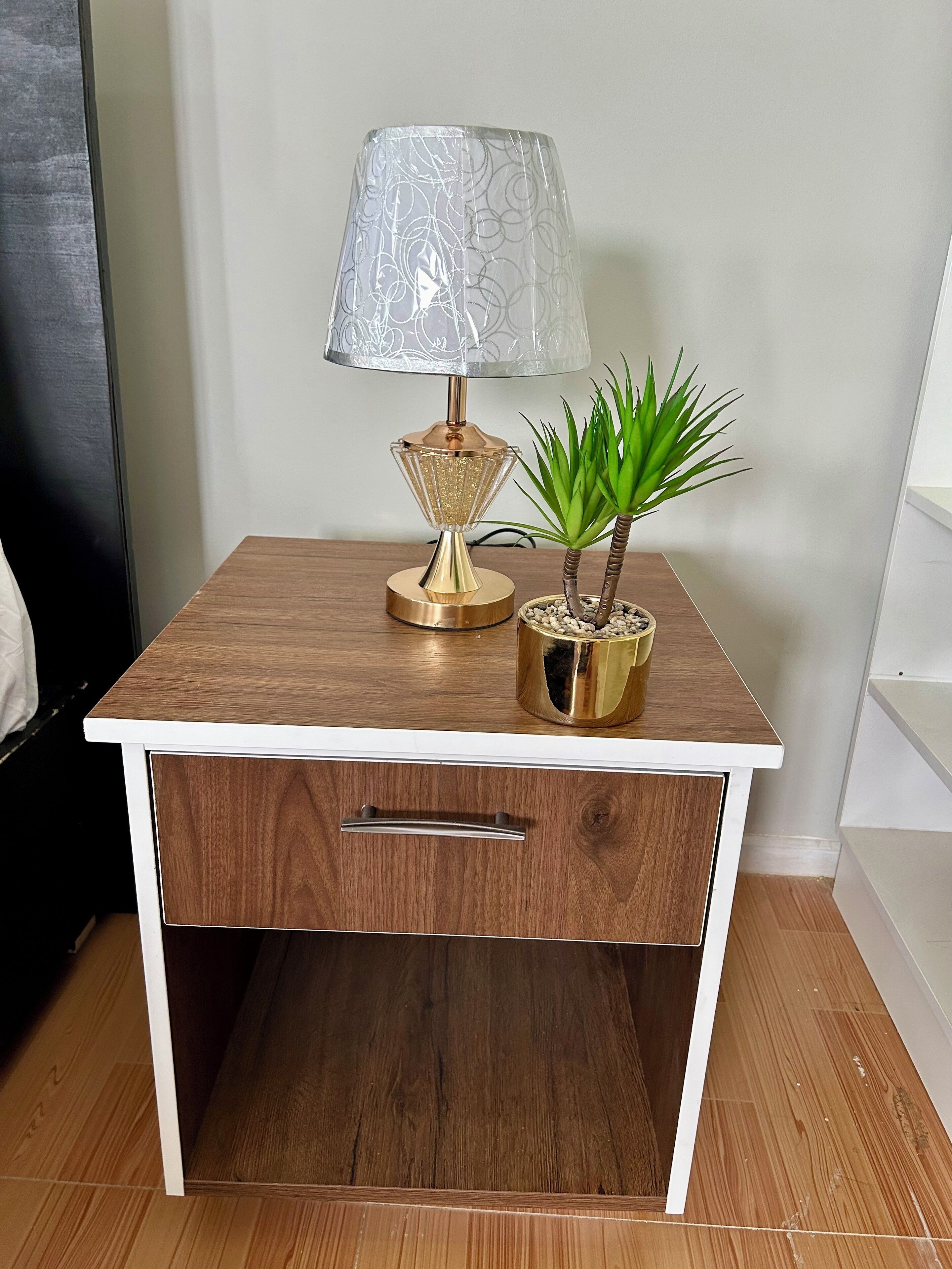 Un pequeño escritorio de madera con un cajón, una lámpara y una planta en maceta.