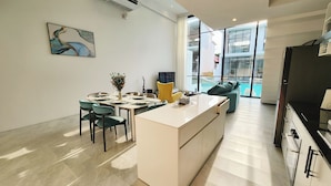 A modern kitchen with a central island, a dining area with chairs and a table, and a large window offering a view of a pool and outdoor area. - Unique Pool Slide Access Townhouse (Bophut)