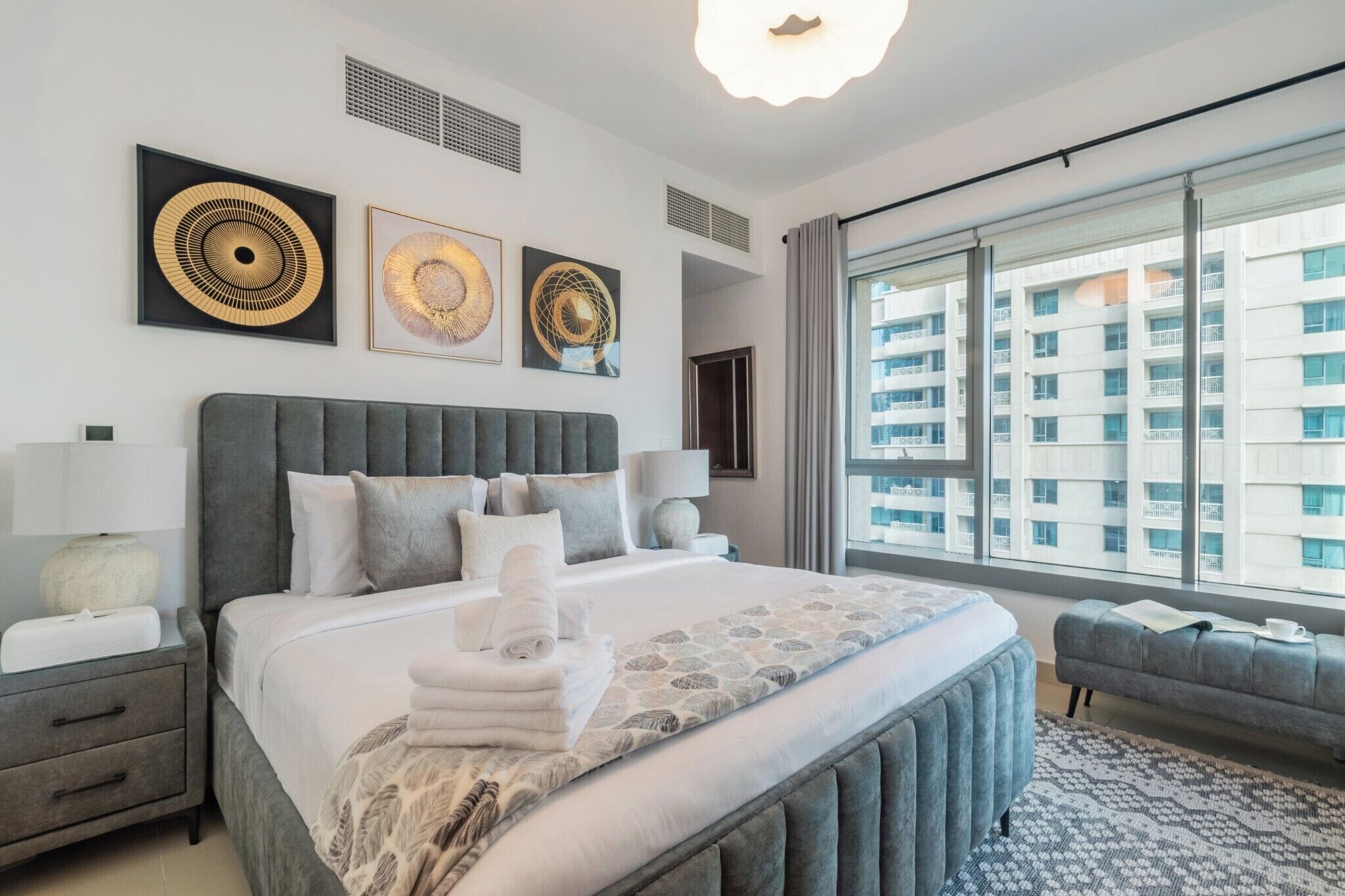 Une chambre d’hôtel moderne avec un grand lit, une tête de lit rembourrée de couleur grise et une vue sur un immeuble de grande hauteur.