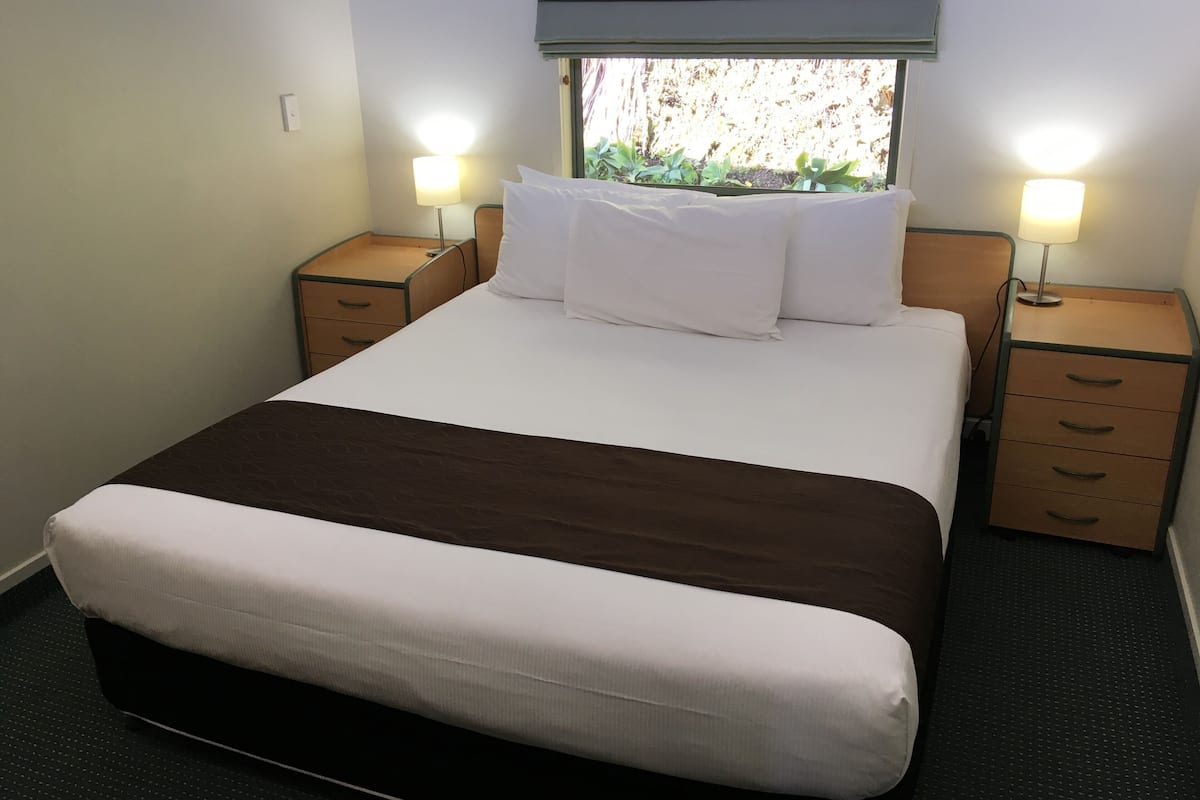 A neatly made bed with white and brown bedding, two bedside tables with lamps, and a window with a view of greenery.