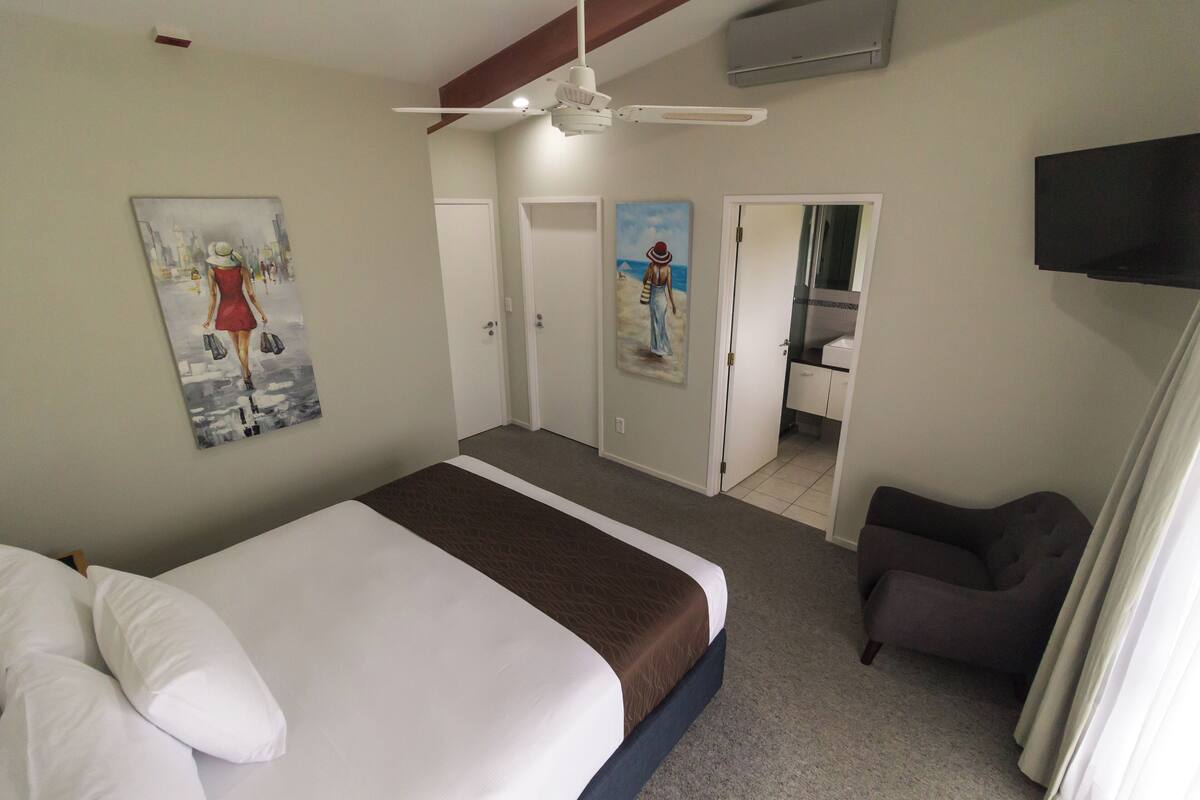 A hotel room with a bed, a ceiling fan, a painting of a woman, a door leading to a bathroom, and a TV mounted on the wall.