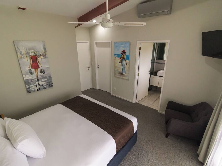 A hotel room with a bed, a ceiling fan, a painting of a woman, a door leading to a bathroom, and a TV mounted on the wall.