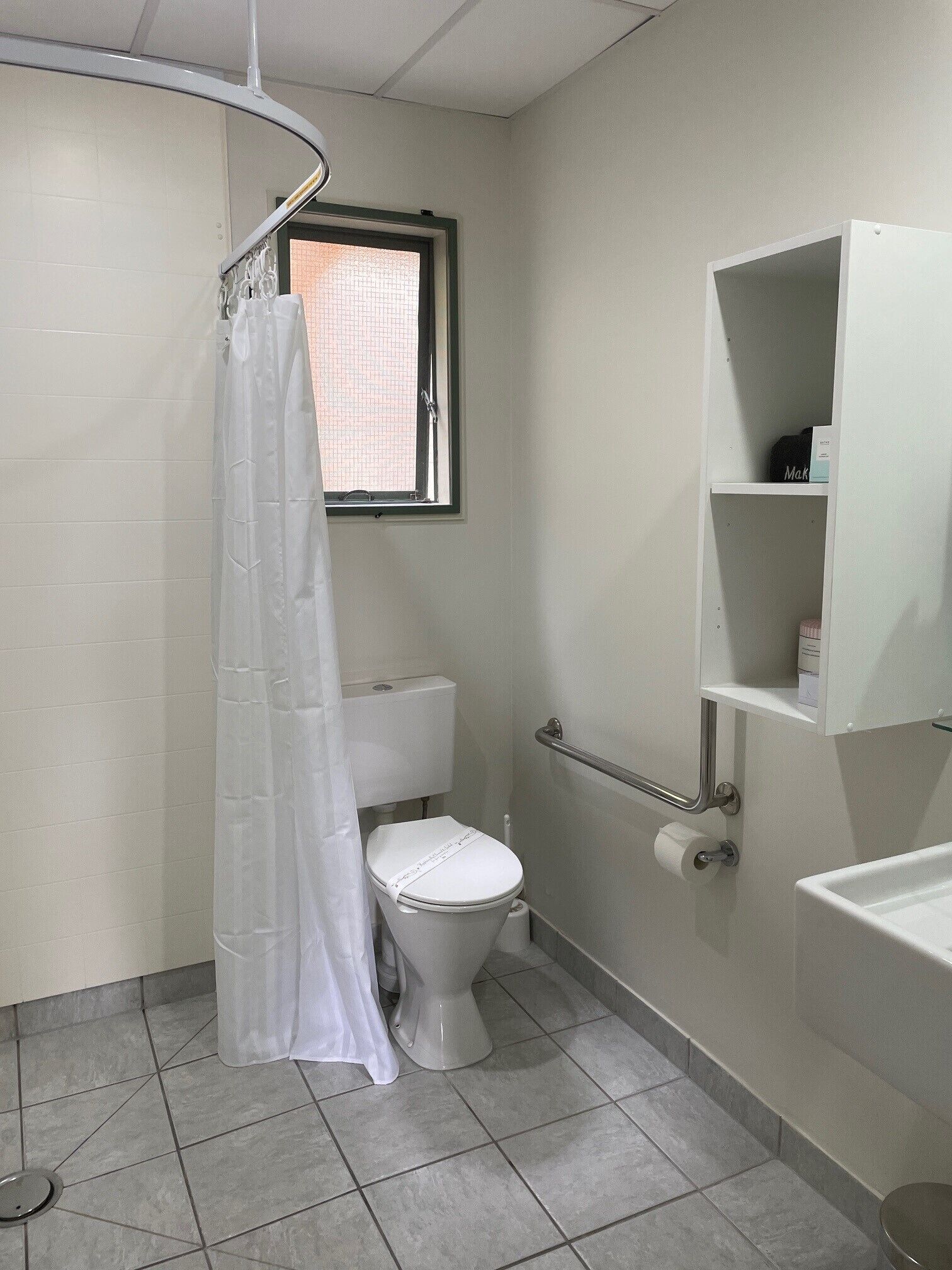 A small, clean bathroom with a toilet, a shower curtain, a window, and a shelf with toiletries.