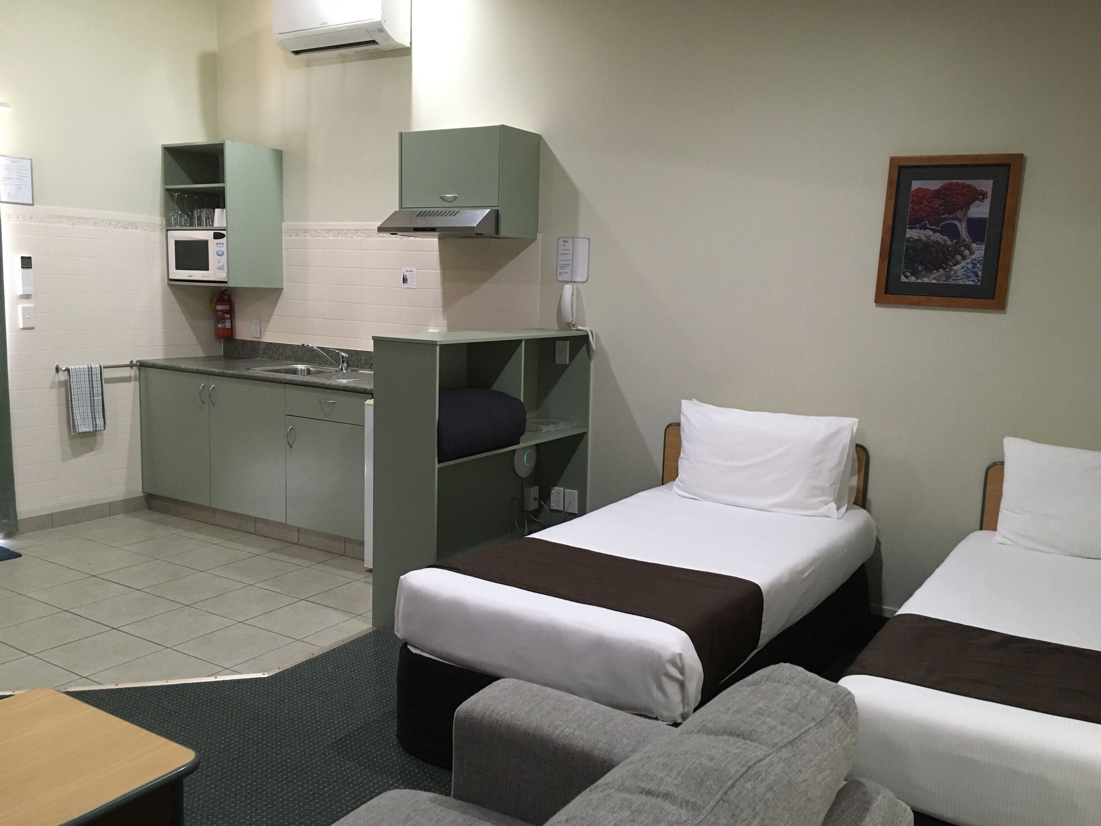 Una habitación de hotel compacta con kitchenette, dos camas individuales, un pequeño sofá y un cuadro en la pared.