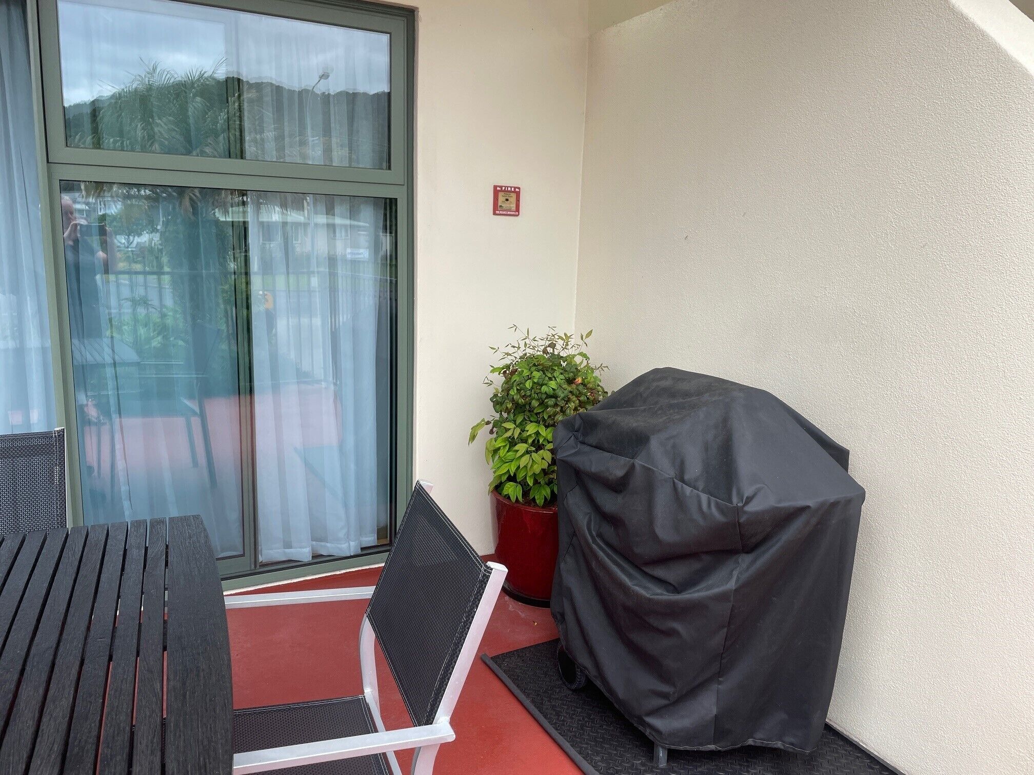 A balcony with a covered grill, a chair, a potted plant, and a window with a fire safety sign.