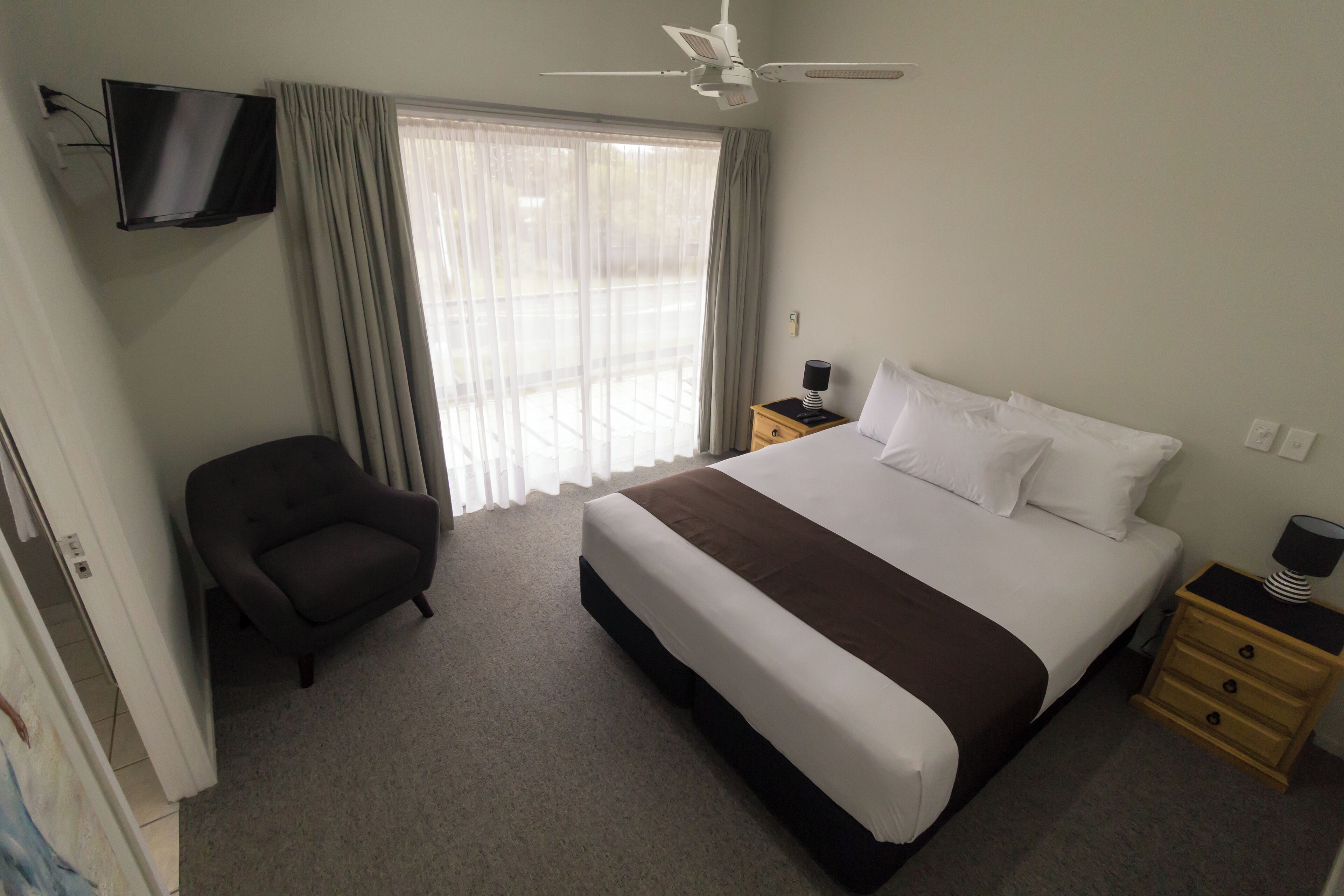 Quarto de hotel com cama, cadeira, televisão fixada na parede, ventilador de teto e janela com cortinas.