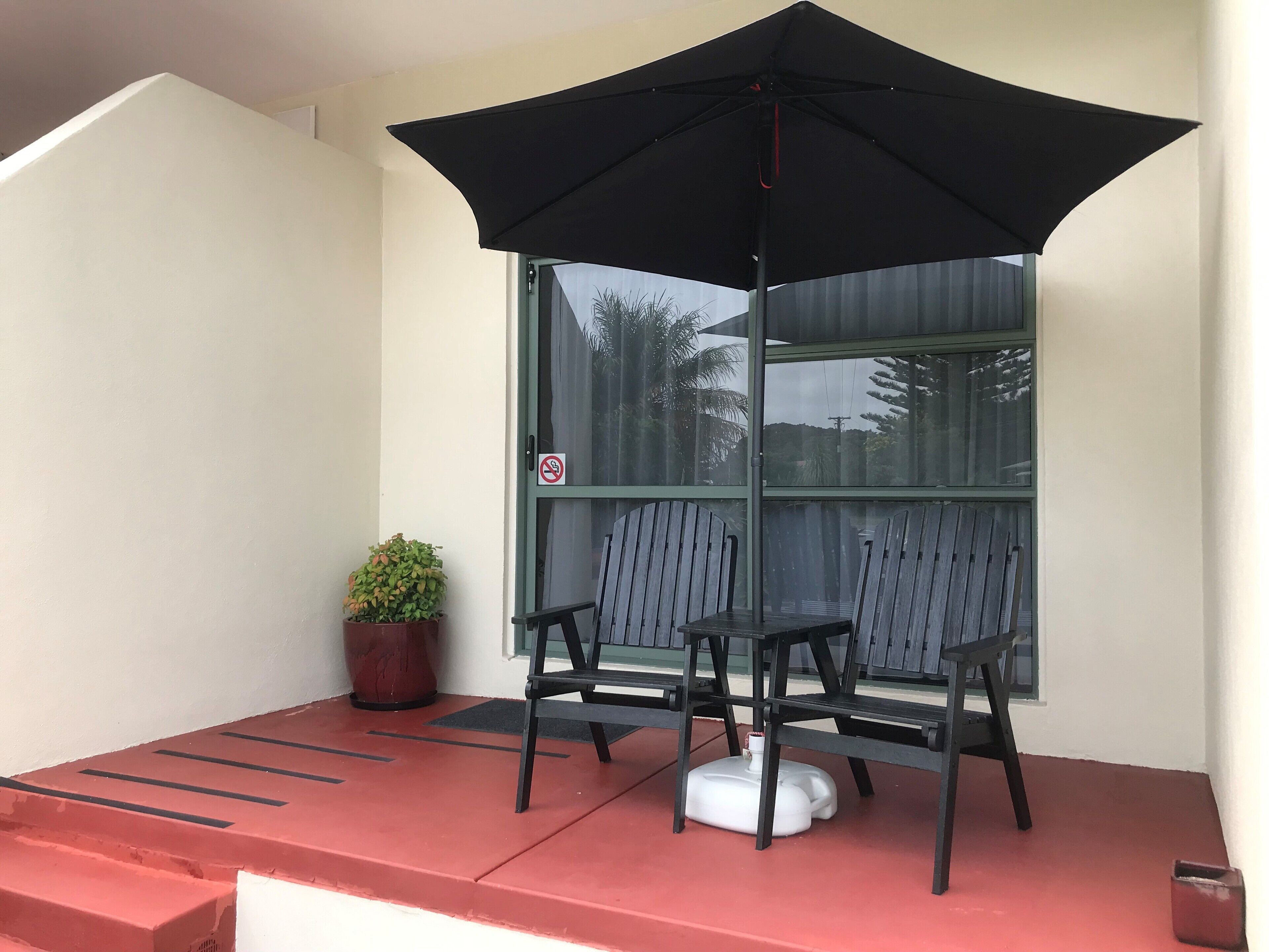 A covered patio with two chairs and a table, a potted plant, and a window with a no smoking sign.